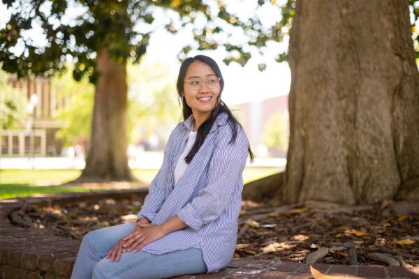 Pa Yang sits on a brick wall in the shade of a giant oak tree in front of Kendall Hall.