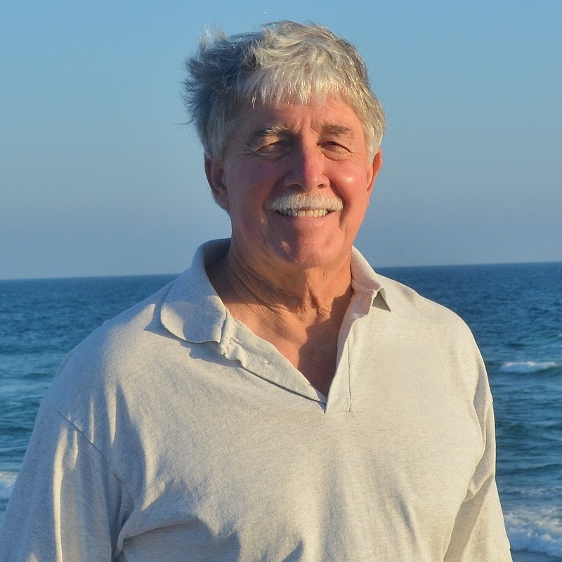 Steven Callan poses for a profile picture wearing a white golf shirt, standing in front of the ocean.