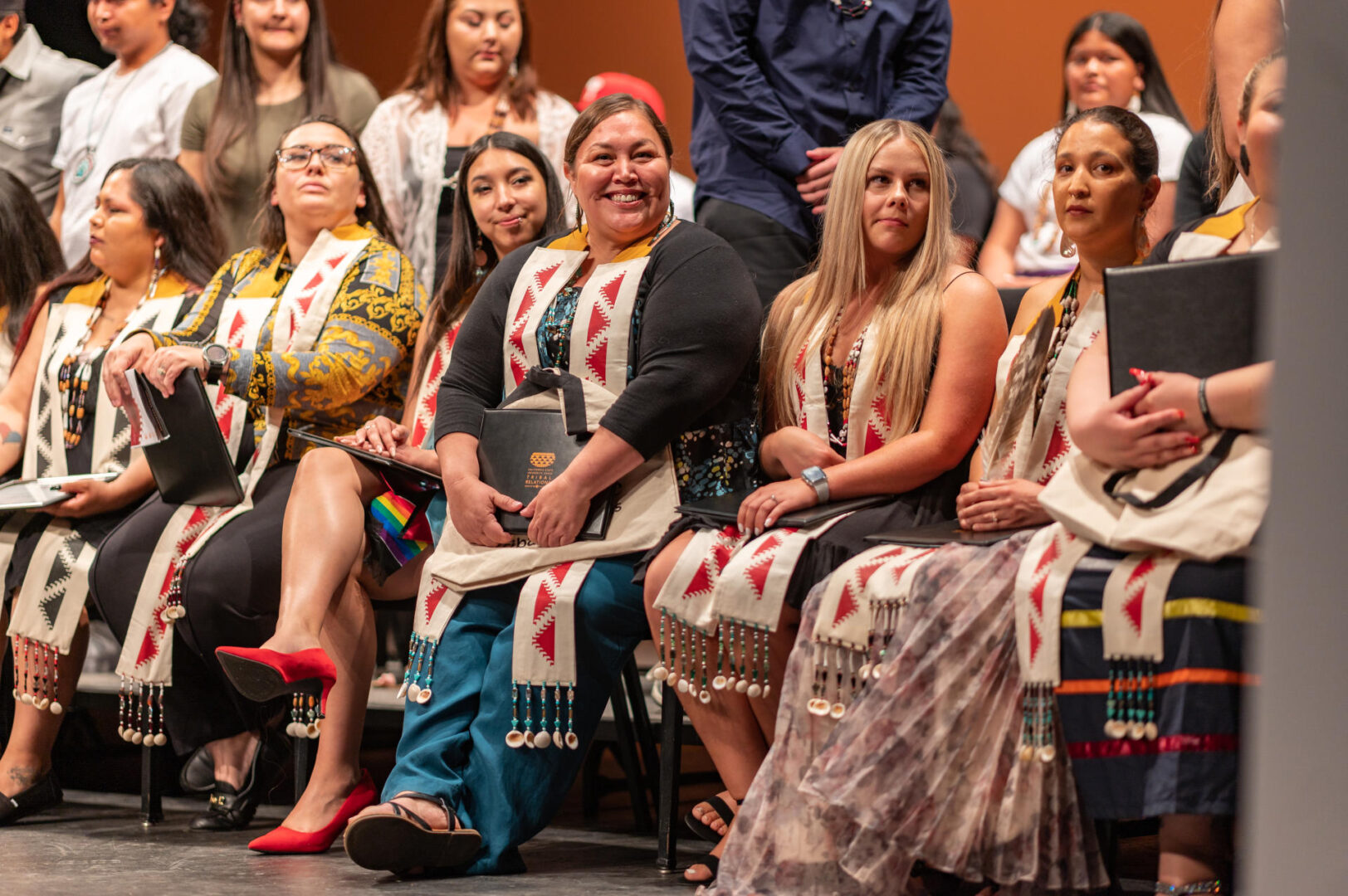 Several Indigenous students sit in chairs smiling during a college graduation