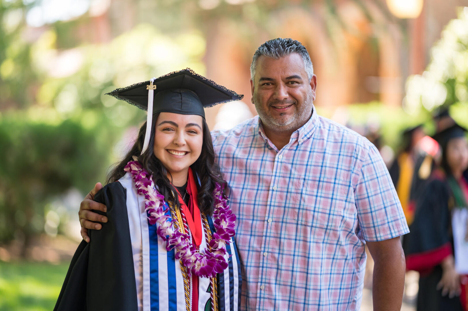 a graduating student wearing cap and gown smiles with a man having his arm around her