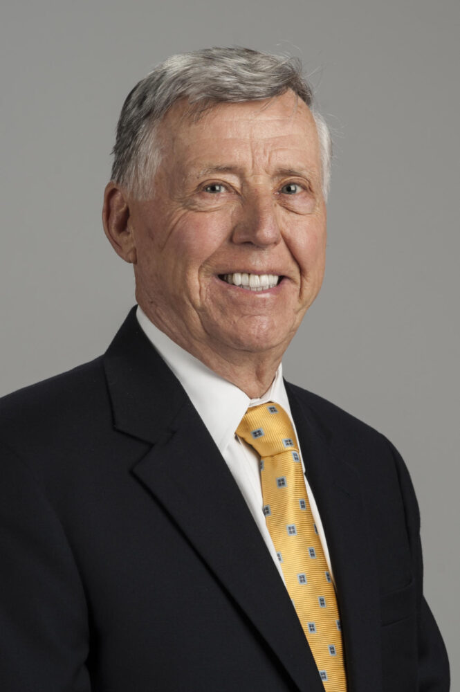 A portrait photograph of Robert Main, who is wearing a dark suit, white dress shirt, and a yellow tie. 