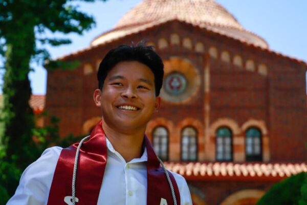 Jonny Nguyen wears a graduation sash and smiles in front of a college building