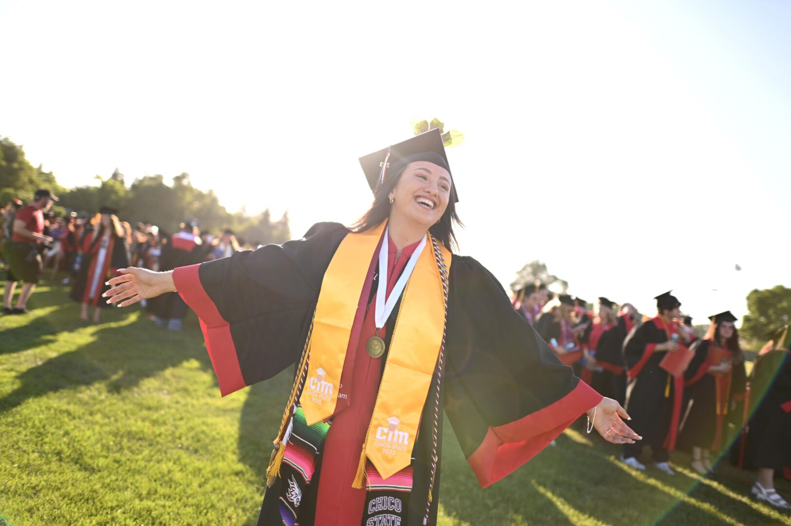 A college student in cap and gown stands smiling with arms wide open