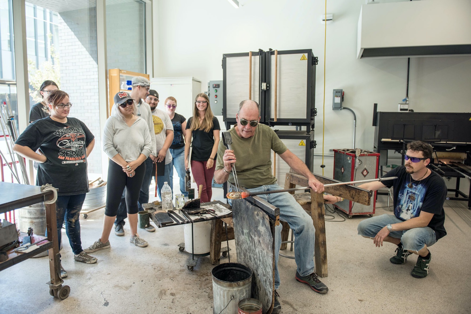 Students stand behind Robert Herhusky as he sits and demonstrates a glassblowing technique