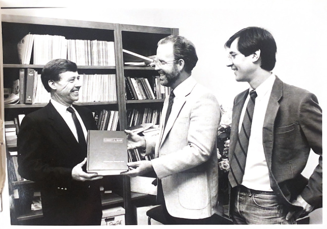 Bob Main receives a book from a second man in the photo. A third man in the photo looks on and smiles. Behind them all are books stacked in a bookcase.