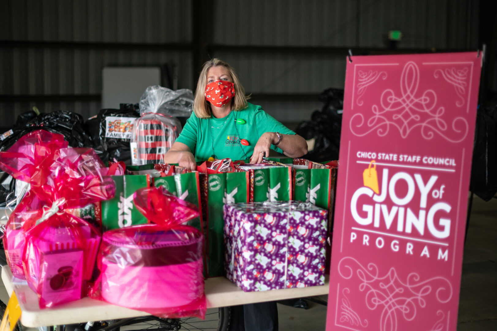 A woman wearing a green t-shirt and face mask stands behind a table full of presents with a large sign that reads, "Chico State Staff Council Joy of Giving Program" in front of the table. 