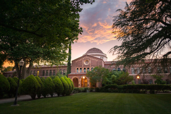 The sun sets behind an academic building with grass and oak trees in the foreground and puffy clouds in the sky