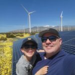Brandon Reinhardt and his young song pose for selfie in front of solar panels and wind turbines with the mountains in the distance.