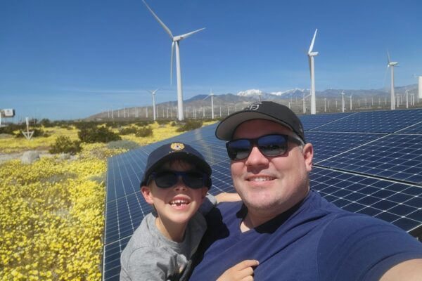 Brandon Reinhardt and his young song pose for selfie in front of solar panels and wind turbines with the mountains in the distance.