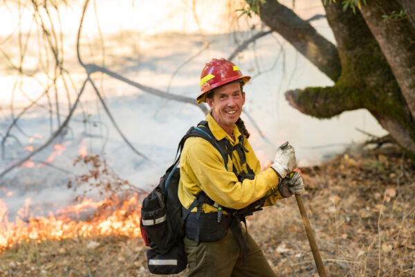Don Hankins leans on a shovel while he smiles and stands beneath a tree during a prescribed fire exercise