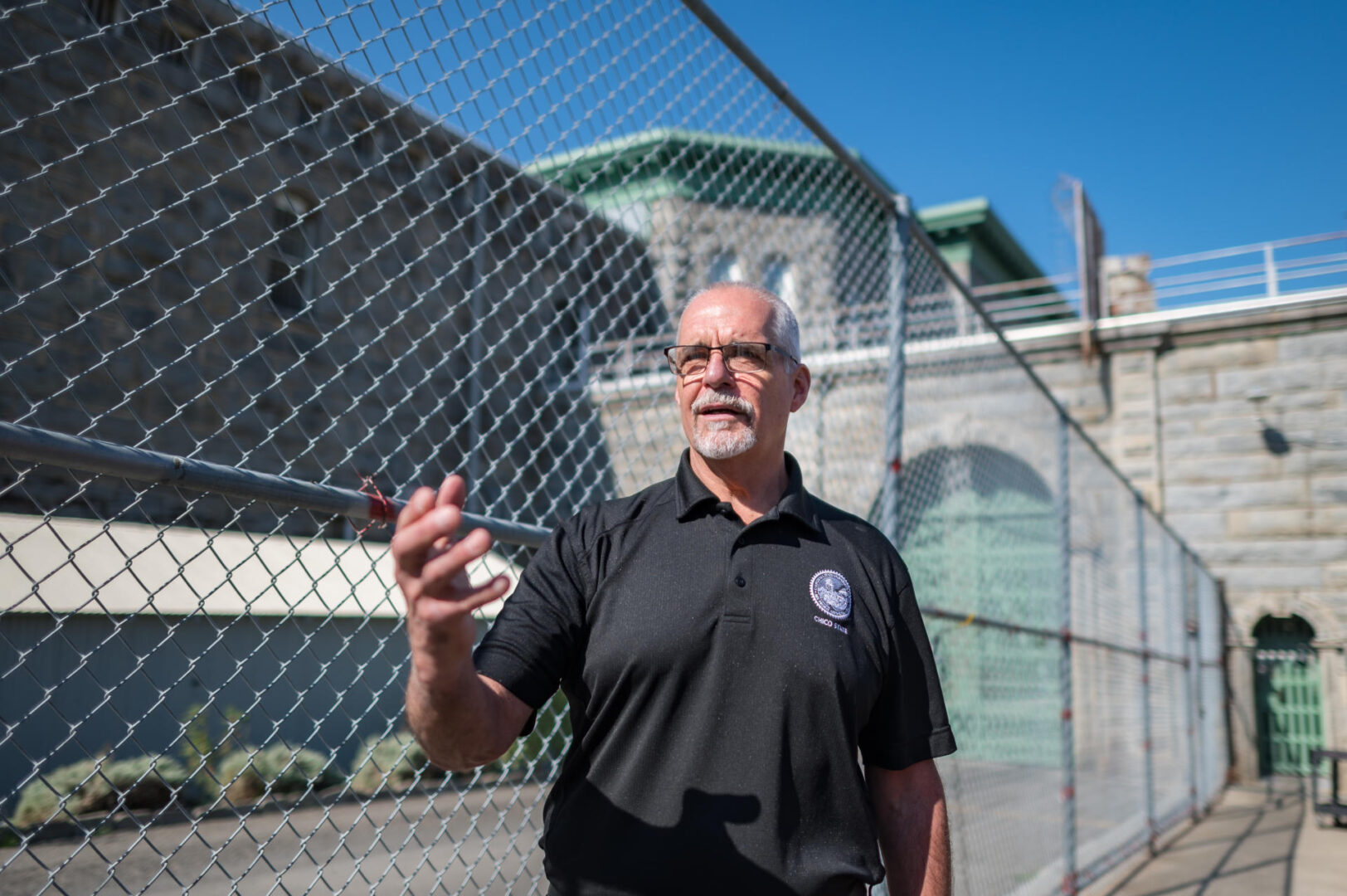 Rory Thelen, wearing a black polo shirt, is photographed during a tour of Folsom prison. He is standing outside with a chain-link fence in the background. 