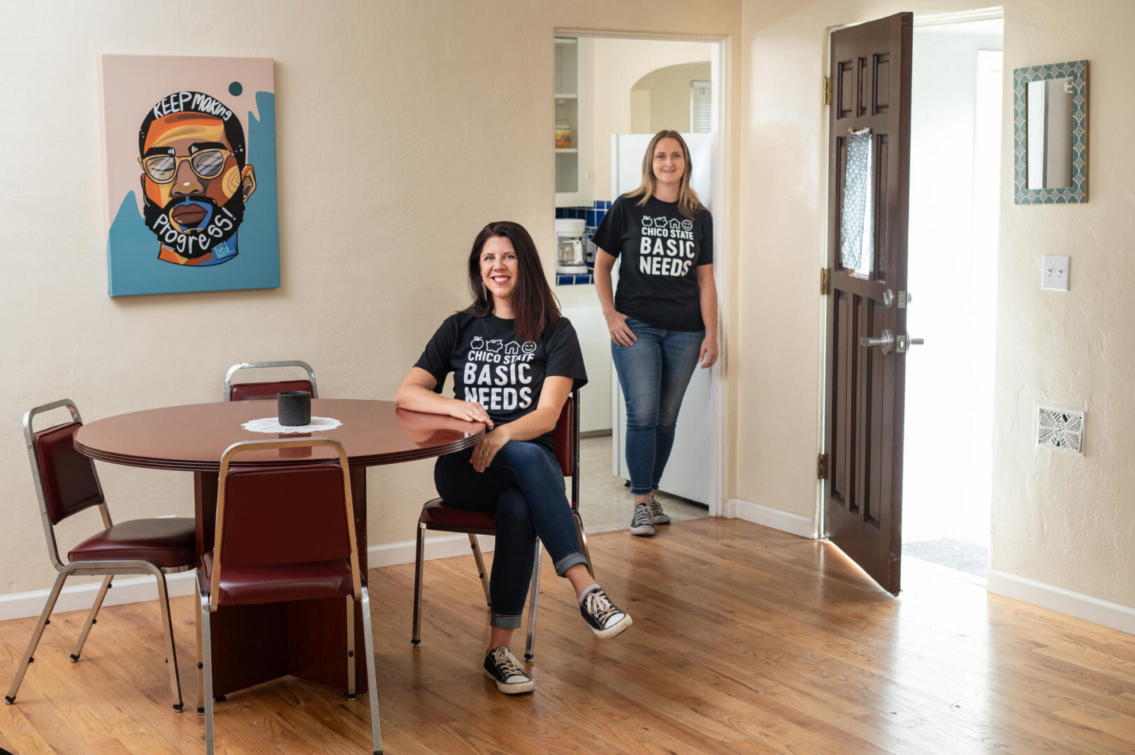 Dawn Carini sits in a chair at a table and Emma Jewett stands in a doorway, both smiling as they pose next to an open front door.