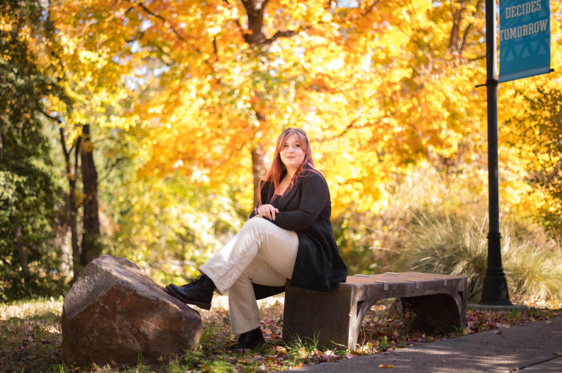 Kate Minderhoud sits on a bench flanked in the golden light of autumn trees.