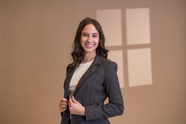 Student Sierra Bowling smiles as she poses with her hands holding her gray jacket in front of a beige backdrop with the outline of a window in the background.