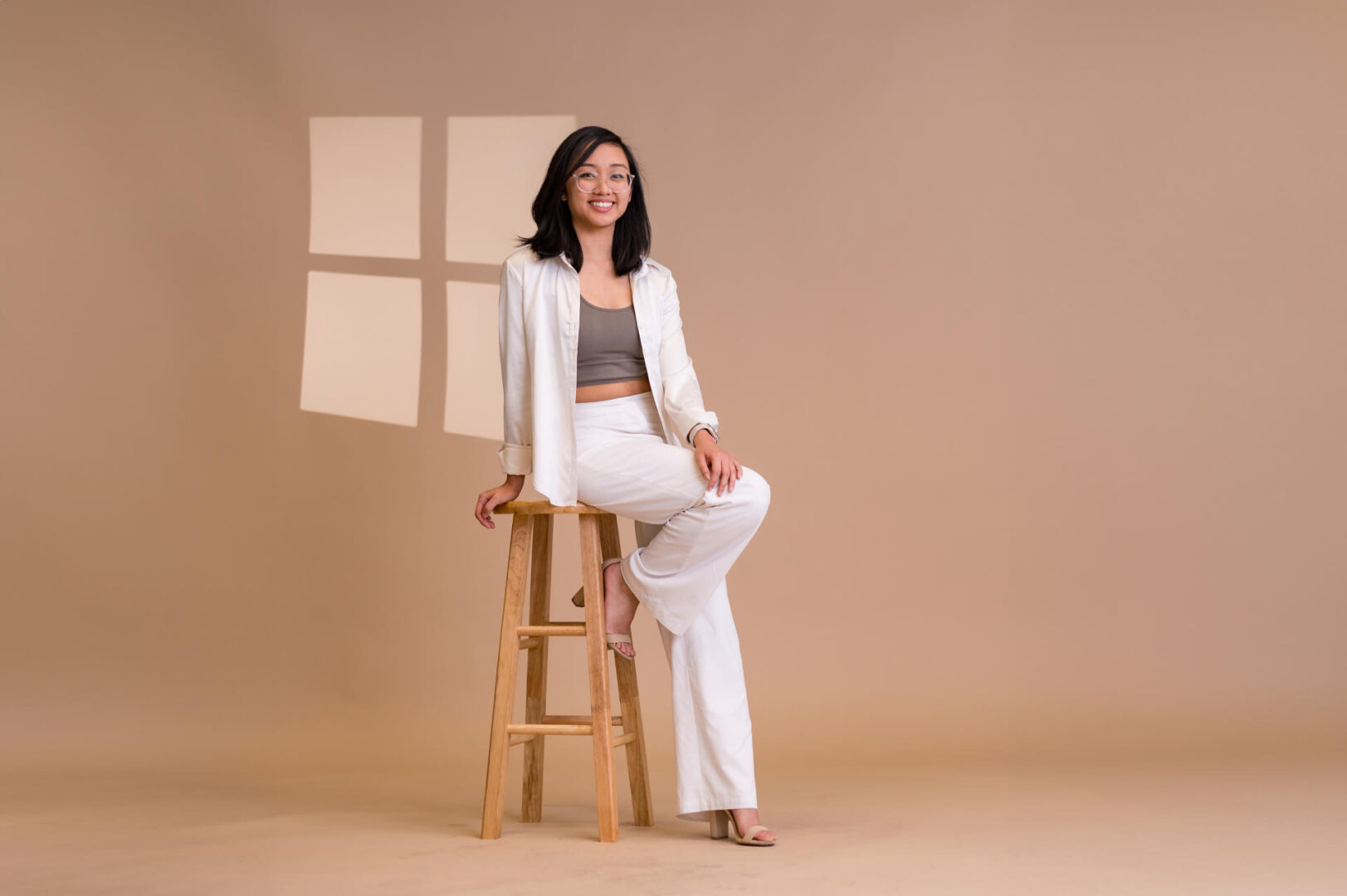 Marifren Francisco, wearing a white suit, sits on a stool with one leg down in front of a neutral background during a photoshoot.
