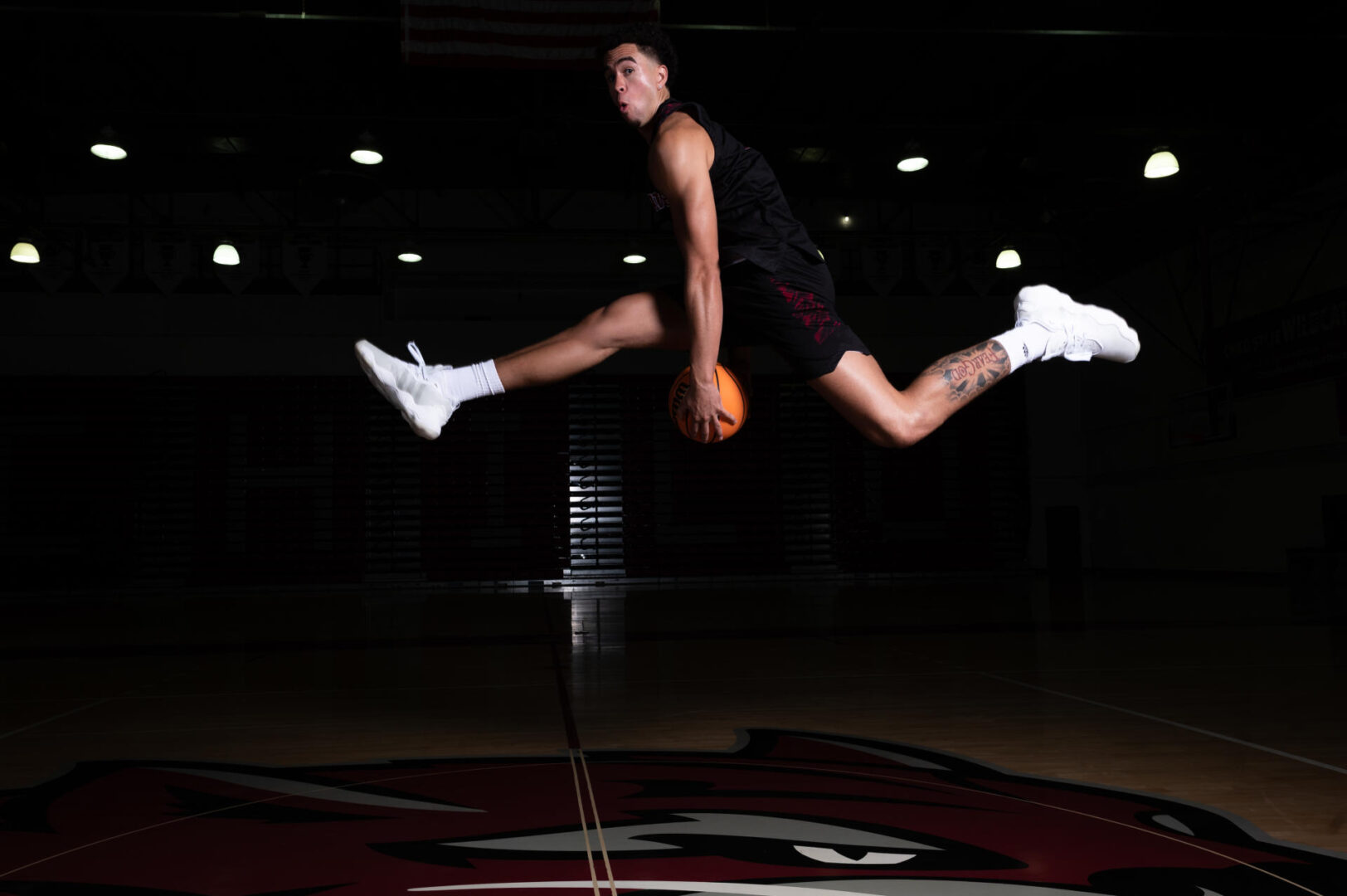 Men’s basketball player Evan Oliver stares at the camera mid-jump in the air with his legs in a near split and a basketball in between his legs.