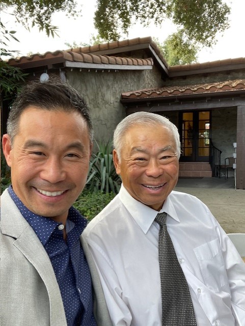 Dave Tu (left) and Norman Tu smile for a photo while standing outdoors in front of a home. 