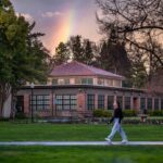A rainbow flashes its colors behind Colusa Hall as a student walks down a sidewalk soaked by recent rains.
