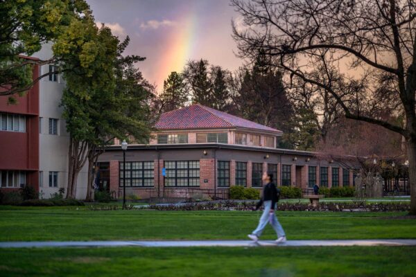 A rainbow flashes its colors behind Colusa Hall as a student walks down a sidewalk soaked by recent rains.