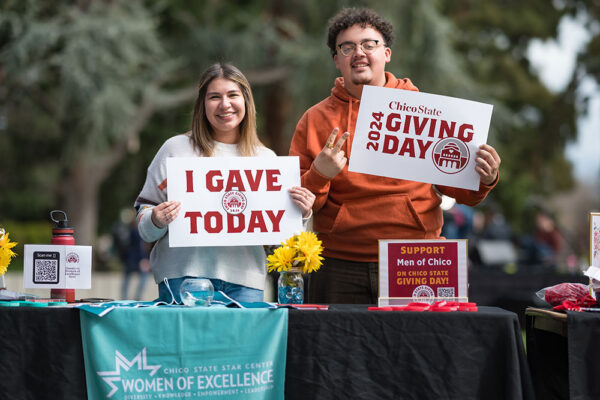 Students Angela Arguelles (left), Christian Hutson (right), representing Women of Excellence and Men of Chico, hold up signs in support of Giving Day 2024 at Chico State.
