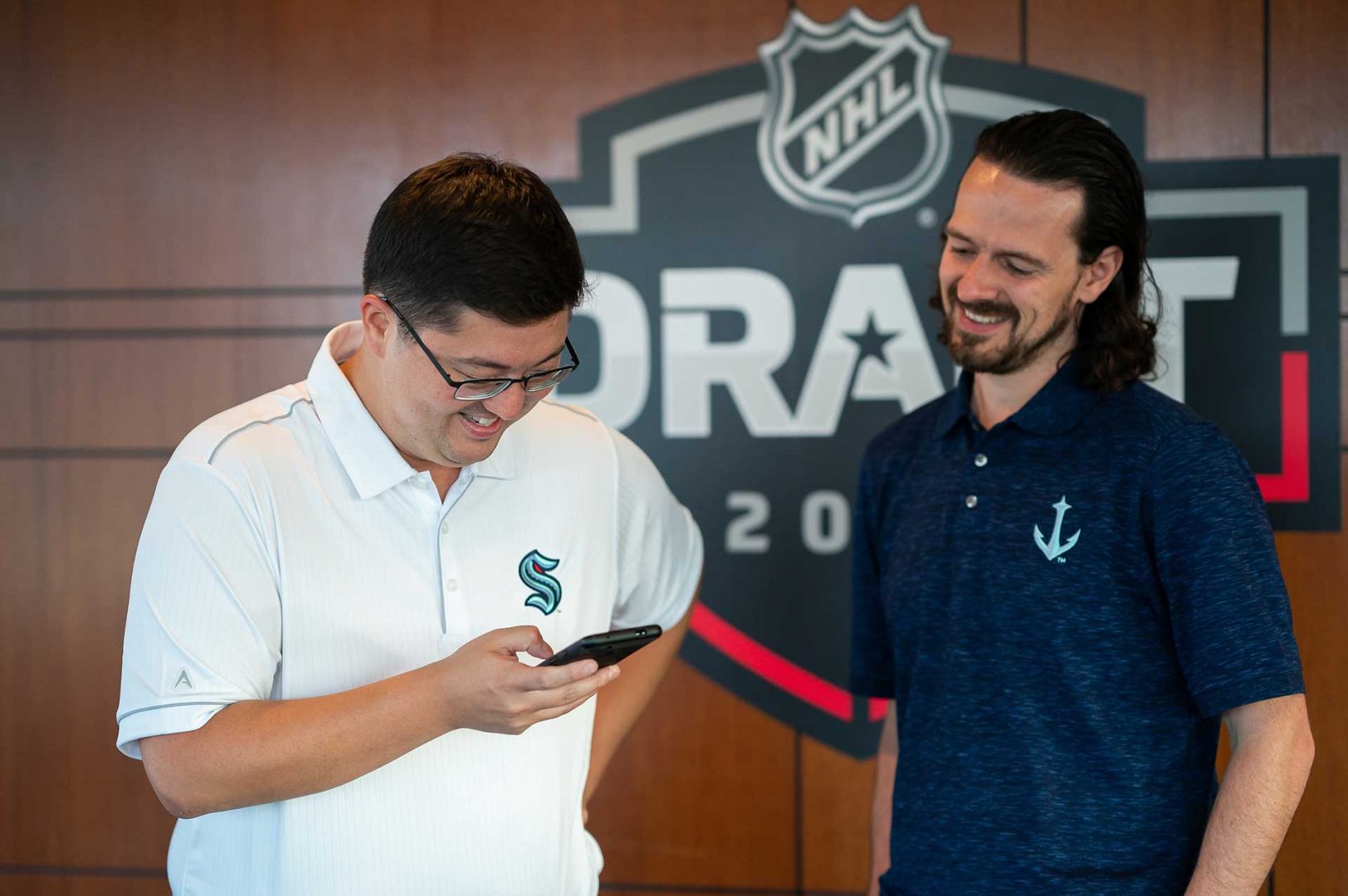 Two men in Seattle Kraken shirts smile as one looks at his phone prior to the NHL expansion draft.