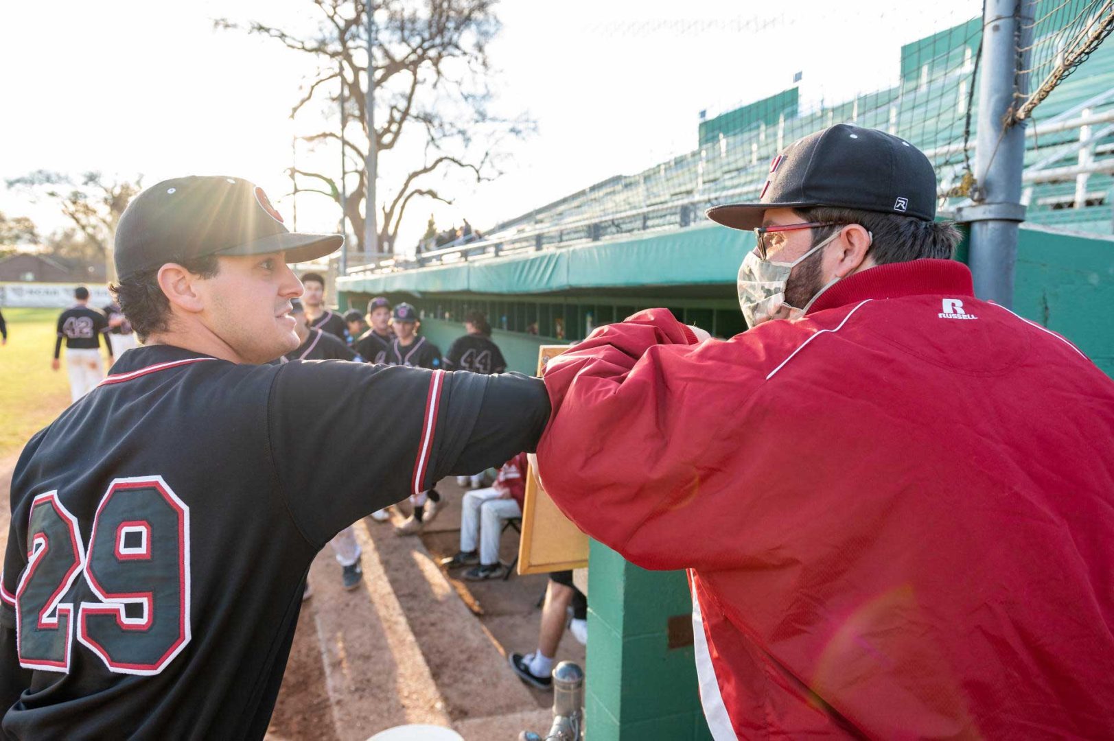 Chico State baseball player Marco Ibarra shares an elbow bump with team superfan John Brownell just outside the Wildcats dugout as the sun sets in the background.