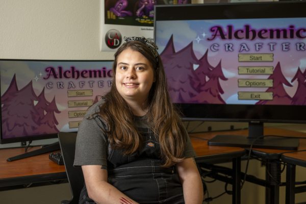 Tyla Chadwick smiles for a portrait as she sits in front of two computer monitors that show the cover of a video game she designed.