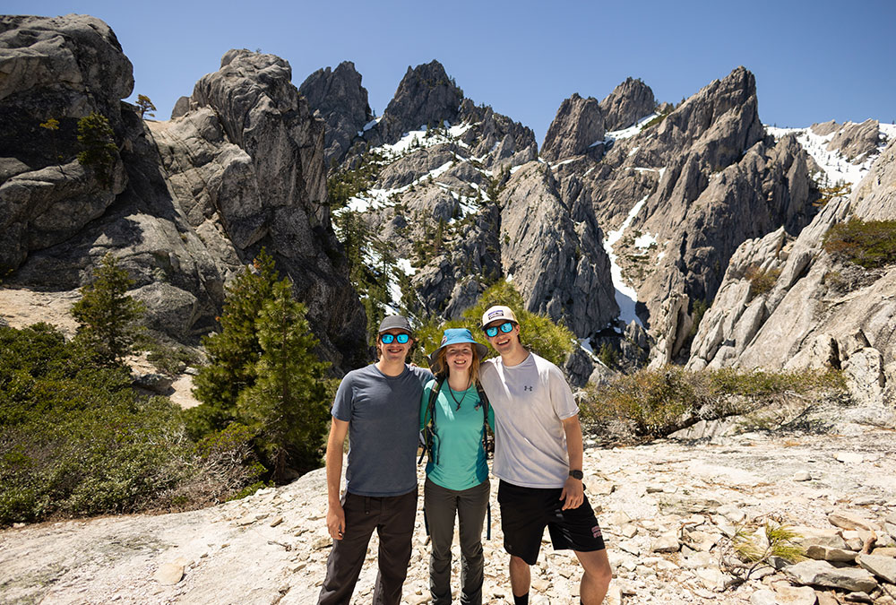 Three hikers stand side-by-side in front of snowy peaks in the mountains.