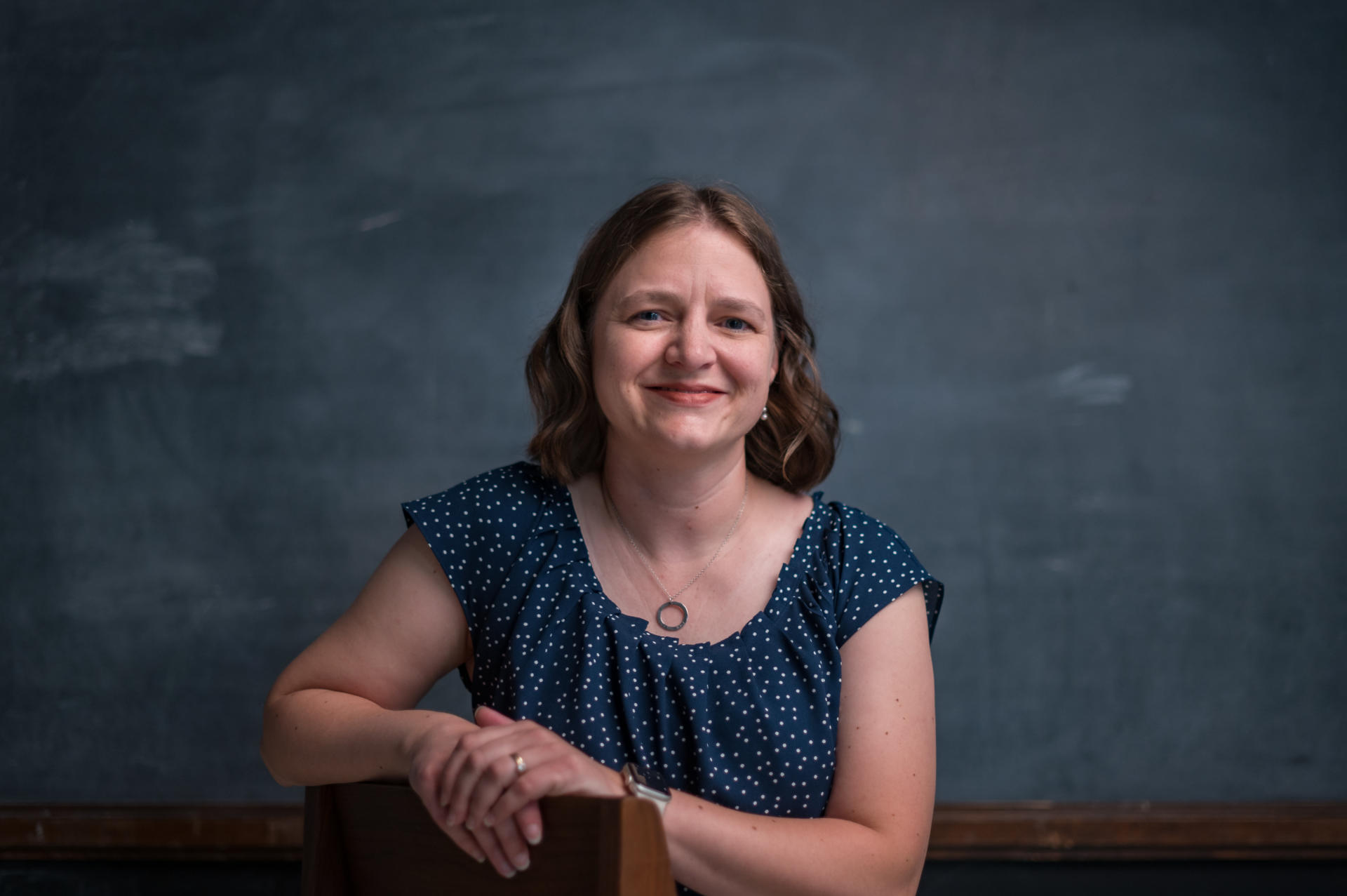 Beth Shook smiles for a portrait in front of a chalkboard.