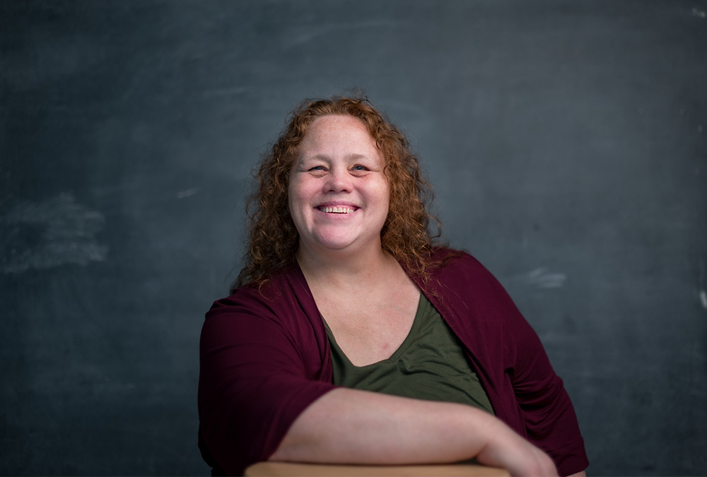 Michell Armeanu is photographed against a chalkboard background, smiling into the camera.