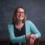 Lecturer Tess Manley is wearing glasses and a light blue sweater, smiling against a chalkboard backdrop.