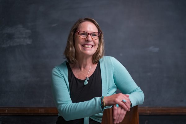 Lecturer Tess Manley is wearing glasses and a light blue sweater, smiling against a chalkboard backdrop.