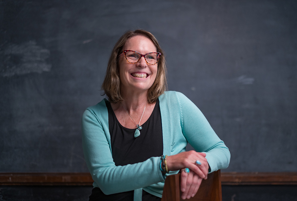 Lecturer Tess Manley is wearing glasses and a light blue sweater, smiling against a chalkboard backdrop.