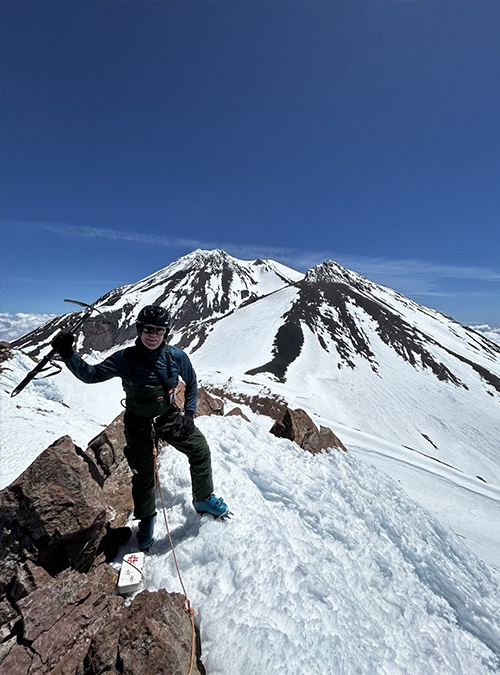 Mark Hendry summits a snowcapped mountain on a sunny day, with his ice pick raised in victory. 