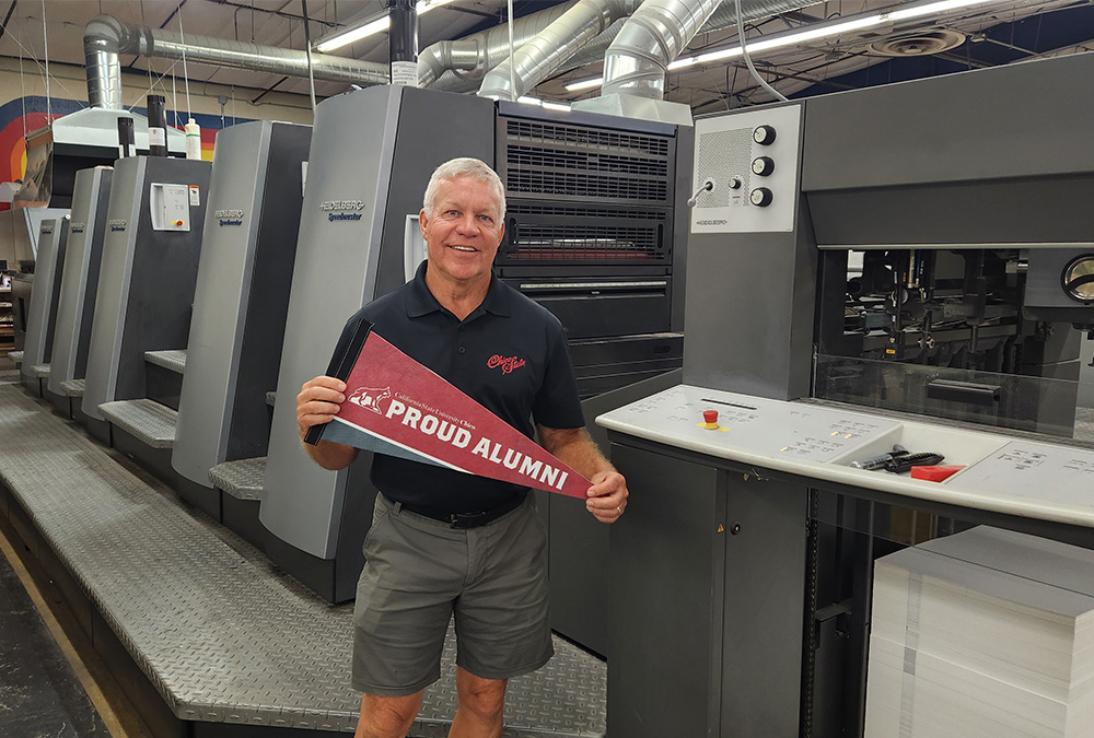 Mark Hendry stands in front of a printing machine holding a red pennant that says "Proud Alumni."